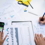 Profissionais colaboram em uma mesa de trabalho, analisando gráficos e documentos com apoio de um celular e um tablet.