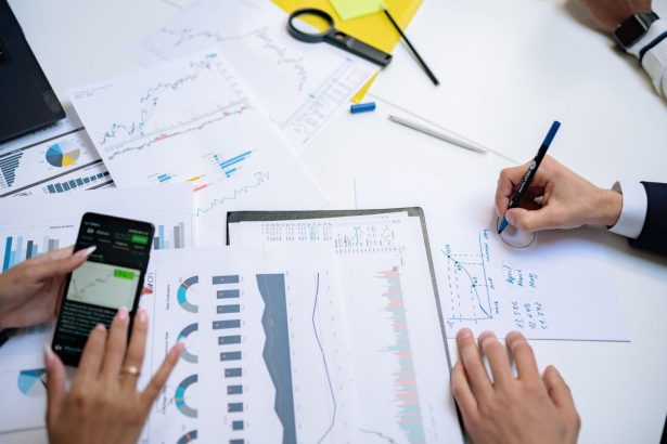 Profissionais colaboram em uma mesa de trabalho, analisando gráficos e documentos com apoio de um celular e um tablet.