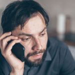 Homem com expressão preocupada usa o celular sentado em um ambiente interno.