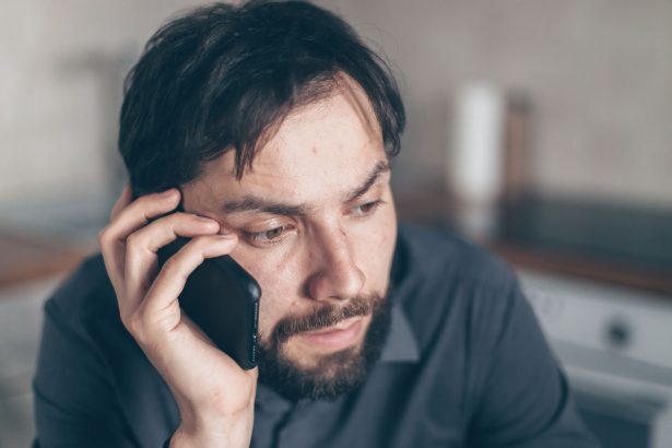 Homem com expressão preocupada usa o celular sentado em um ambiente interno.