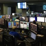 Profissionais trabalham diante de vários monitores em uma sala de controle.